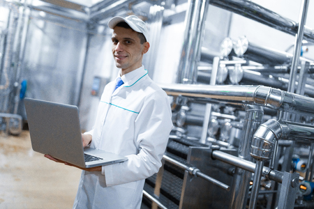 Technologist in a white coat with a laptop in his hands controls the production process in the dairy shop. Quality control at the dairy plantの写真素材