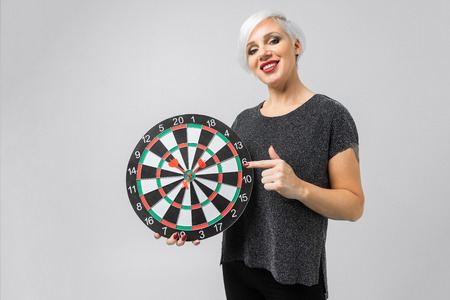 charming blonde holds a target in her hand and points her finger at her in isolation in a light Studio. Business woman smiling and holding big target of darts. Success concept portraitの写真素材