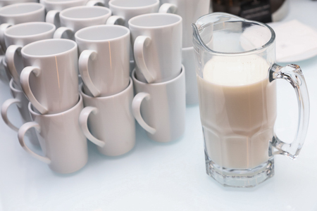 glass jug with white liquid and empty white tea cups stand on the table. Beer glass with milk and pyramid of cupsの写真素材