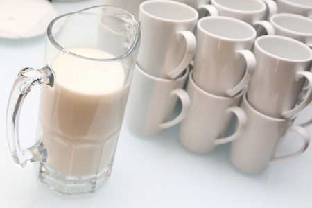 glass jug with white liquid and empty white tea cups stand on the table. Beer glass with milk and pyramid of cupsの写真素材
