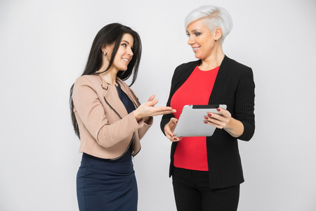 two confident slender buisness ladies holding a tablet. Vertical portrait against wall in studioの写真素材