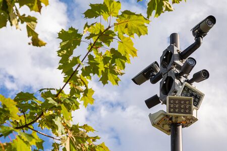 Black surveillance videocameras and white street lamps on one column against sky and green leaves of maple treeの写真素材