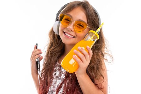 Smiling girl wearing glittery dress and vibrant yellow sunglasses, with big white earphones, listening to music and holding thin cellphone and bottle with orange juice isolatedの写真素材