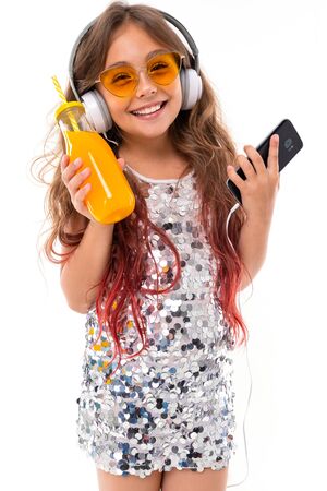 Teenage girl in sparkling dress and vibrant yellow sunglasses, with big white earphones, listening...