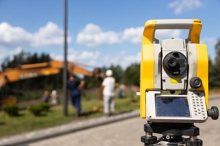 Close-up of yellow and orange tacheometer standing on street against work on construction site....