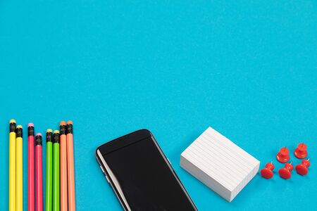 A half visible multicolored pencils, black smart phone, a pile of white scratch paper and a group of...
