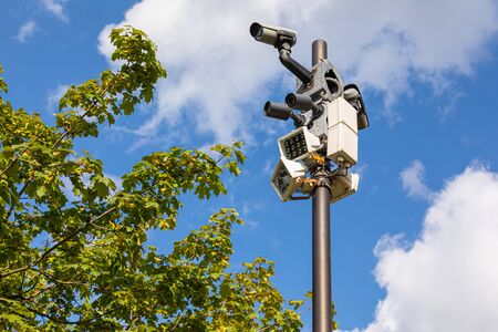 Safety videocameras and white street lamps on one column against sky and green leaves of maple tree