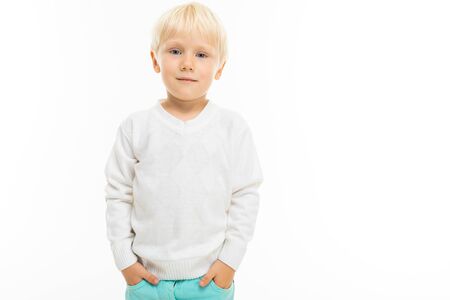 Little boy with short blonde hair, blue eyes, cute appearance, in white jacket, light blue pants, standing with his hands in his pockets.の写真素材