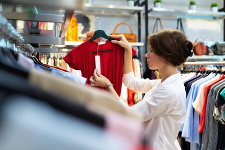 Beautiful caucasian woman chooses red dress, looks price and thinks about her choice.
