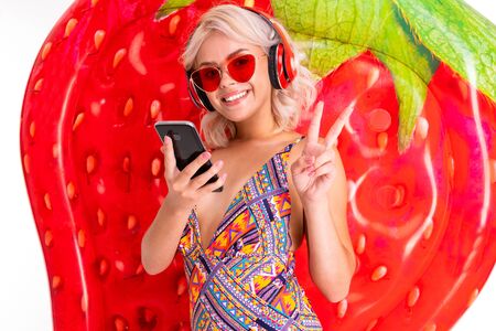 Happy blonde caucasian female stands in swimsuit with big rubber mattress, phone, earphones, do selfie and smiles isolated on white background.の写真素材