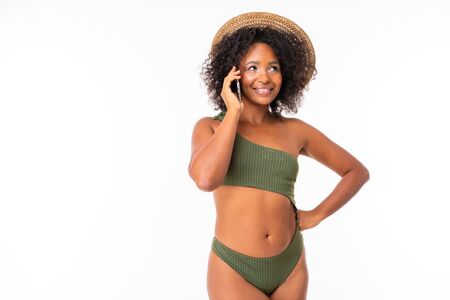 Pretty african female stands in swimsuit, talking on the phone with friends or family and smiles isolated on white background.の写真素材