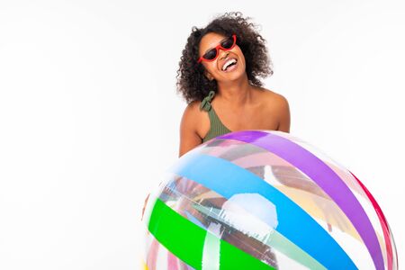 Pretty african female in swimsuit stands with big colourful rubber ball and smiles isolated on white background.の写真素材