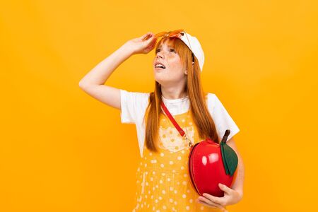 red-haired girl in glasses and with a red handbag in the shape of an apple is grimacing at the camera on a yellow banner background..の写真素材