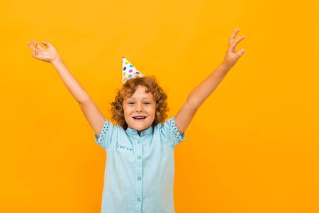 European red-haired curly boy with a festive cap on his head raises his hands up on a yellow background.の写真素材