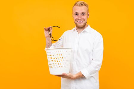 blond man dressed in a classic white shirt throws glasses in the trash bin on a yellow background.の写真素材