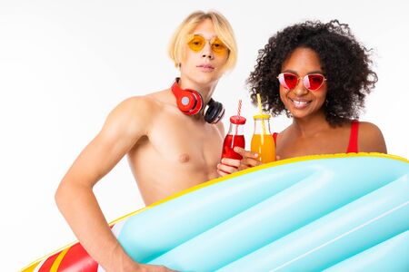 Pretty african female and caucasian blonde man stands in swimsuit with rubber beach mattresses,...