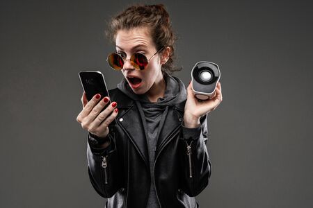 Caucasian girl with rough facial features in a black jacket listen to music with phone and little music column isolated on black background.の写真素材