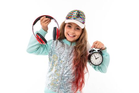 Little pretty caucasian girl in a tracksuit with big earphones and alrm clock, picture isolated on...