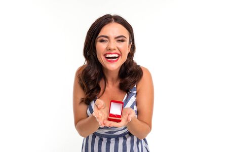 happy girl with brown hair in a dress with a neckline holds out a box with an engagement ring in...