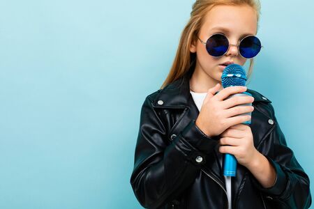 attractive european girl in a leather jacket in sunglasses singing with a microphone on a light blue...