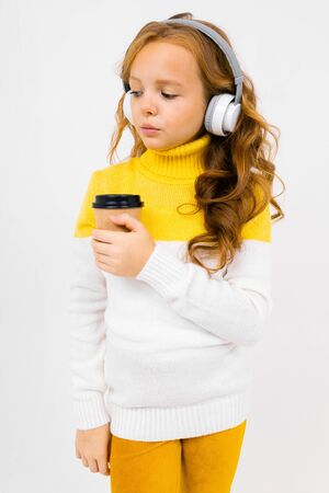 Beautiful teenager caucasian girl with yellow trousers, white hoody listen to music with big earphones and drinks coffee isolated on white backgroundの写真素材