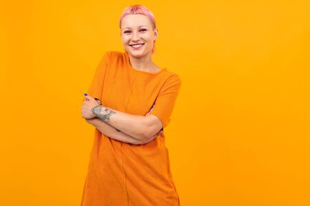 Happy caucasian pretty woman with short pink hair, tattoo in orange dress smiles isolated on orange...
