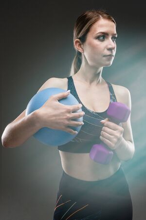 slim girl in sportswear holds a sports ball and a dumbbell on a black background with lens flare...