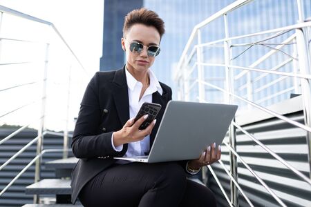 Serious caucasian woman in black and white office clothes do freelance with laptop and waits colleague near the office buildingの写真素材