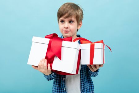 Caucasian boy holds many white boxes with gifts and rejoices, portrait isolated on blue background.の写真素材