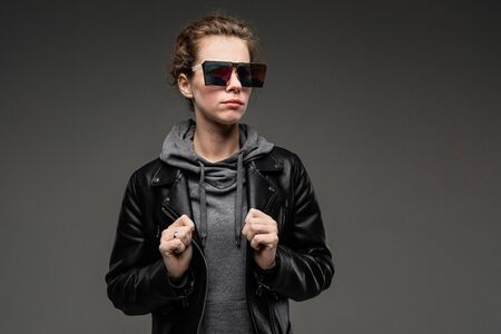 attractive caucasian girl in stylish glasses, a gray sweatshirt and a black leather jacket stands on...