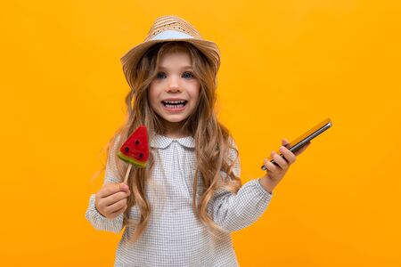 cute little smiling girl with a lollipop and a phone in her hands on a yellow background with copy...