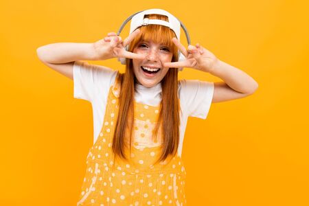 Happy girl with red hair listen to music with big white earphones and smiles with isolated on yellow...