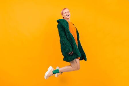 Extraordinary beautiful woman with short pink hair in dark coat smiles and jumps isolated on orange...