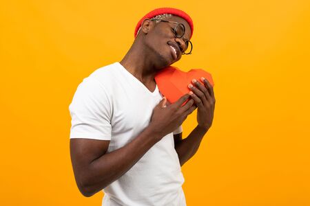 stylish african man with a beautiful snow-white smile hugs a red heart made of paper for Valentine's day on a yellow studio background.の写真素材
