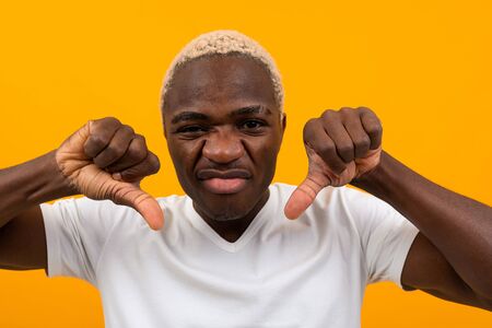 portrait of a blond displeased charismatic african man in a white t-shirt holding his fingers down on a yellow studio background.の写真素材