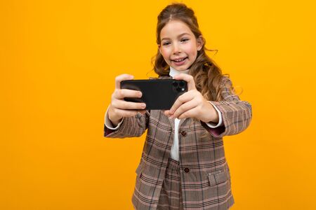laughing stylish blonde caucasian girl in a classic jacket takes a photo on a smartphone on a yellow studio background.の写真素材