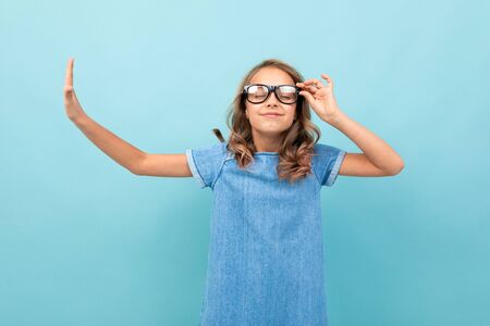 Schoolgirl with wavy brown hair in blue dress with eyeglasses smiles isolated on blue background.の写真素材