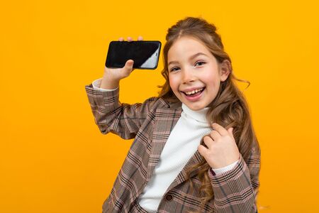 cute smiling young girl in a classic brown jacket shows a phone with a mockup on a yellow background.の写真素材