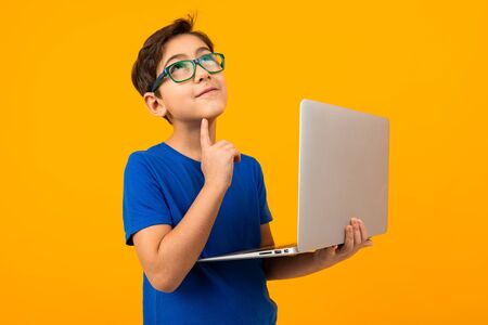 smart boy in a blue t-shirt thinking holding a laptop in his hands on a yellow studio background.の写真素材