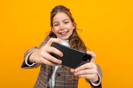 laughing stylish blonde girl in a classic jacket plays games on a smartphone on a yellow studio background.の写真素材