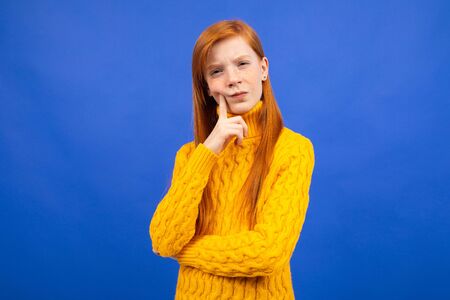 attractive caucasian red-haired girl in a yellow sweater stands thoughtfully on a blue studio...
