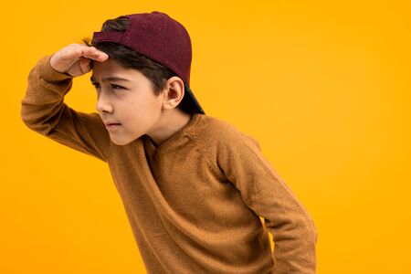 European handsome guy teenager in a brown sweater and baseball cap is looking for a target on a yellow background.の写真素材