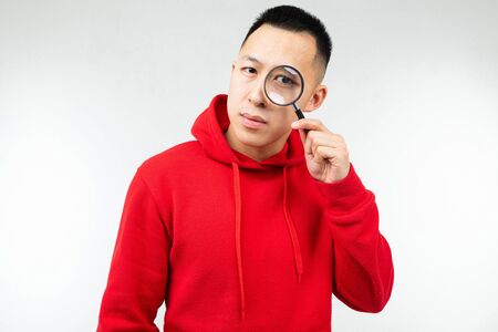 brunette man in red hoody looks at the camera through a magnifier on a white isolated background.