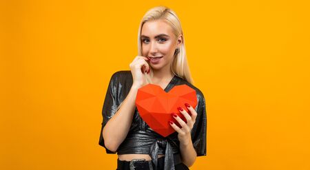 European girl holding a 3D heart figure in her hands on an orange studio background. Valentines Dayの写真素材