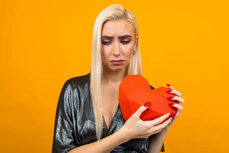 sad european young woman holding a red heart in her hands on an orange studio background