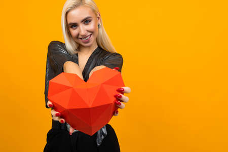 cute blond girl holds out a 3D heart figure on a yellow background with copy space