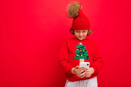 a young man with a smartphone in his hands against the background of a red wall, in a knitted hat and a sweater with a Christmas treeの写真素材