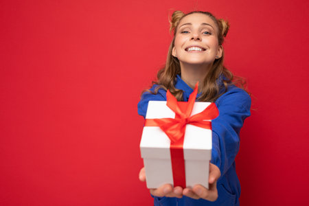 Shot of attractive happy smiling young blonde woman isolated over red background wall wearing blue trendy hoodie holding white gift box and looking at camera. Empty spaceの写真素材