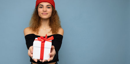 Shot of beautiful positive smiling young dark blonde curly woman isolated over blue background wall wearing black crop top and red hat holding gift box looking at camera. Free spaceの写真素材