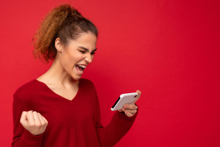 Young emotional woman wearing dark red sweater isolated over red background holding smartphone having fun and showing yes gesture looking at mobile phone screen.の写真素材
